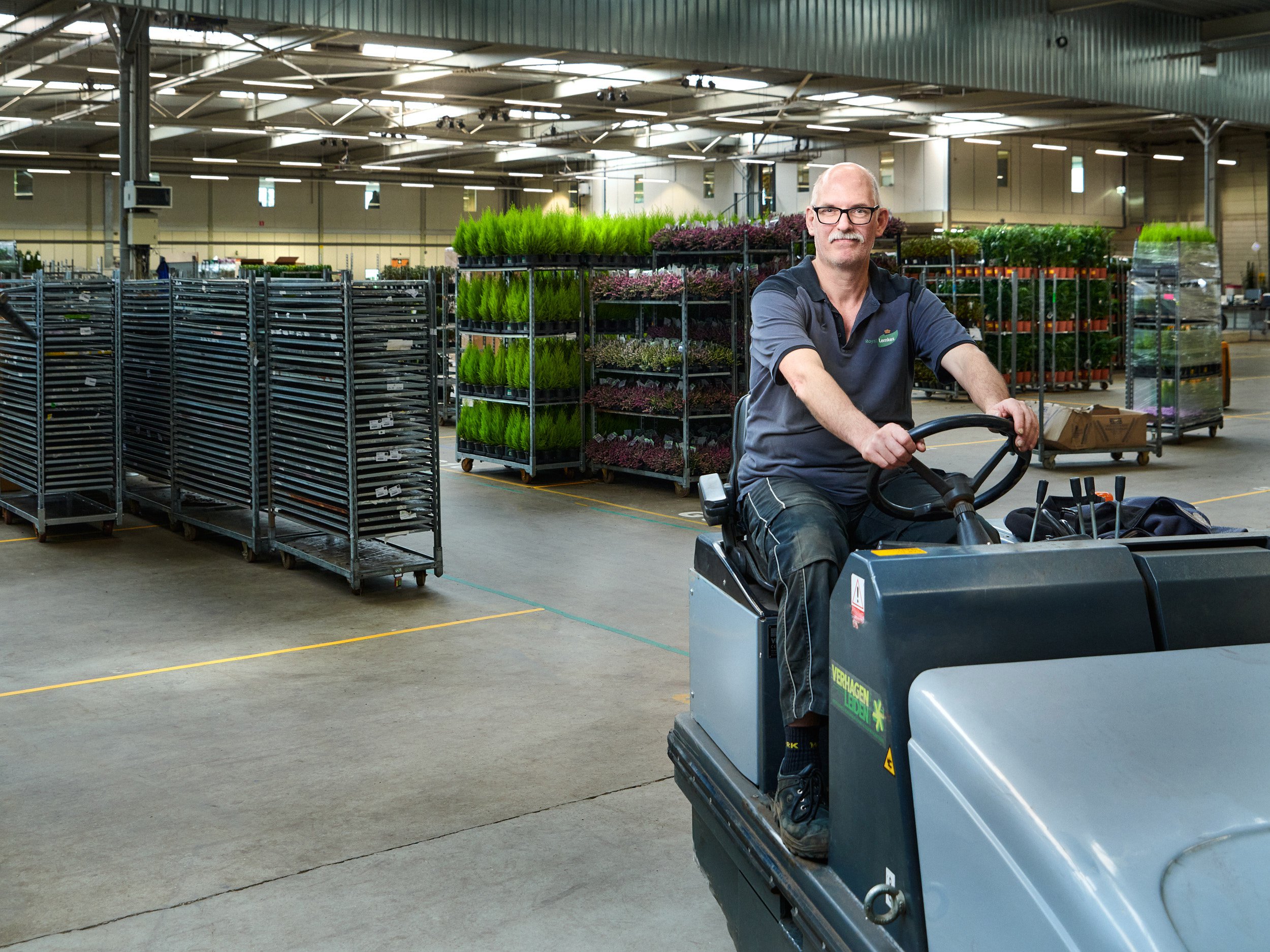 Medewerker rijdt op een schoonmaakmachine in een kas vol planten en karren.